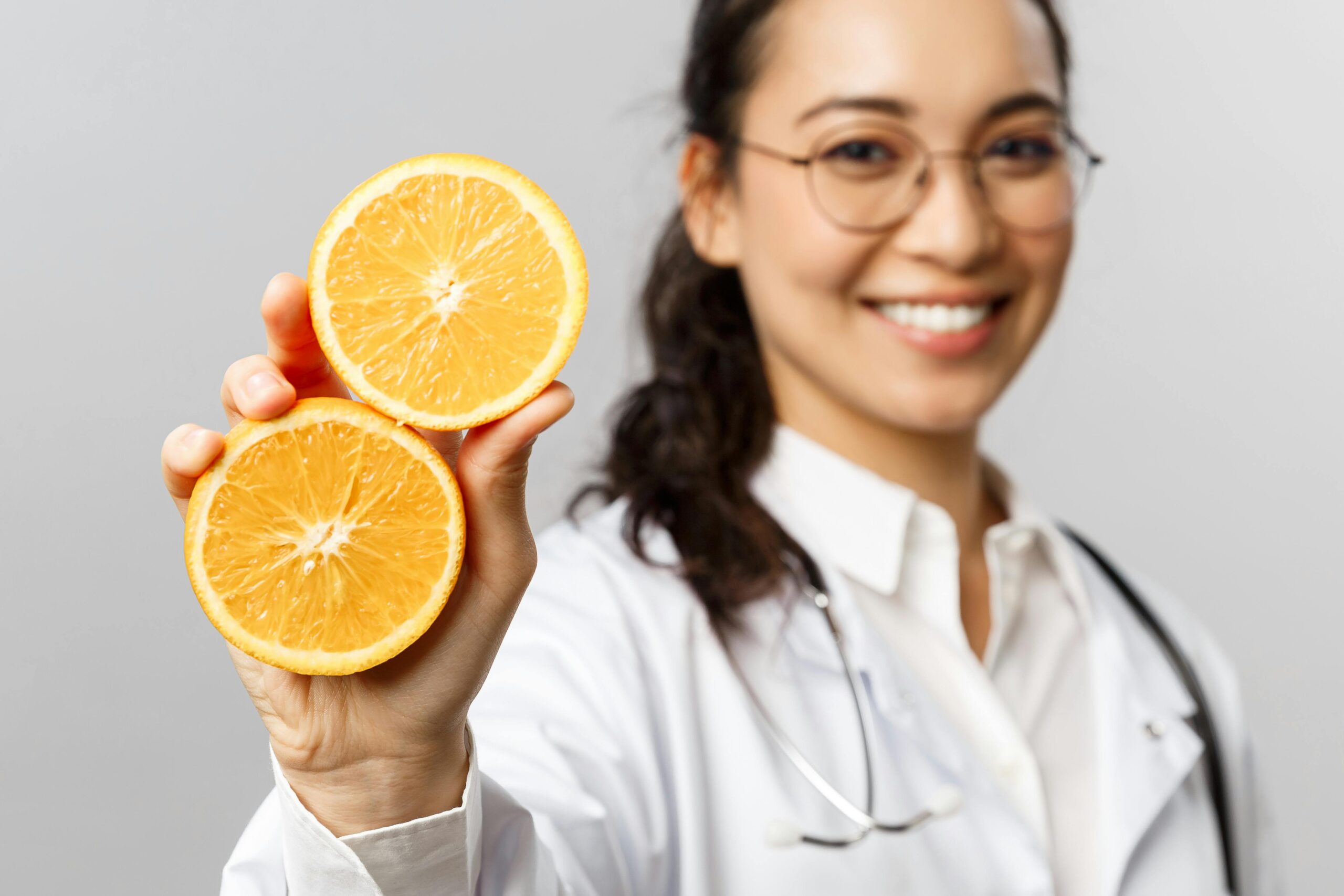 Cheerful female doctor with glasses presenting two halves of a sliced orange, promoting health.